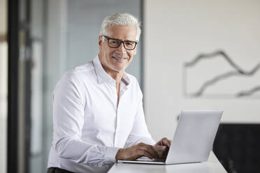 Älterer Geschäftsmann lächelt, während er im Büro vor einem Laptop steht - RBF08111