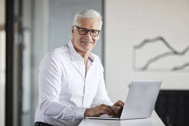 Mature businessman smiling while standing by laptop in office - RBF08111