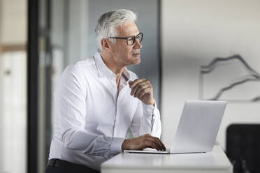 Businessman with laptop looking away in office - RBF08113