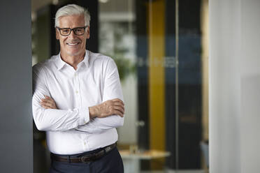 Businessman with arms crossed leaning on wall in office - RBF08147