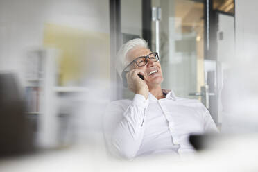 Männlicher Unternehmer, der lächelt, während er im Büro mit einem Mobiltelefon spricht - RBF08159