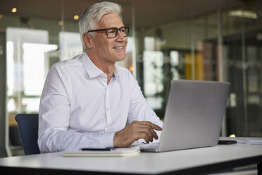 Smiling businessman with laptop contemplating in office - RBF08174