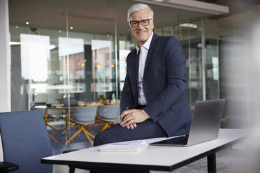 Mature male professional sitting on desk by laptop in office - RBF08180