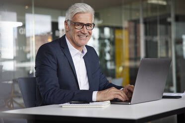 Lächelnder Geschäftsmann mit Laptop am Schreibtisch im Büro - RBF08182
