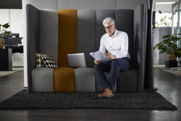 Männlicher Unternehmer liest ein Dokument, während er im Büro am Laptop sitzt - RBF08197
