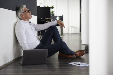 Tired businessman sitting by laptop on floor in office - RBF08199