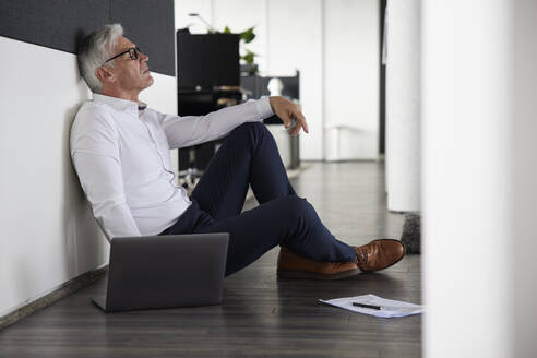 Müder Geschäftsmann sitzt mit Laptop auf dem Boden im Büro - RBF08199