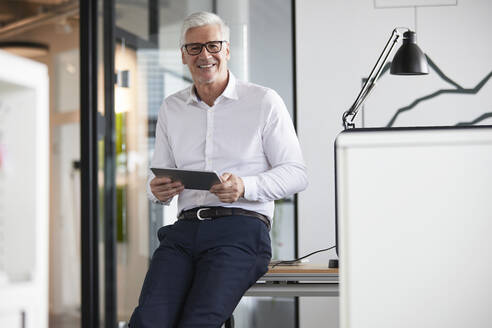 Lächelnder Unternehmer mit digitalem Tablet, der sich auf einen Tisch im Büro stützt - RBF08203