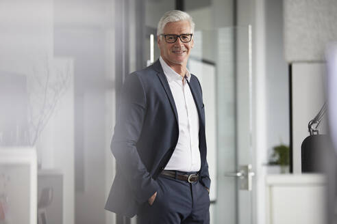 Smiling businessman with hands in pockets standing at office - RBF08211