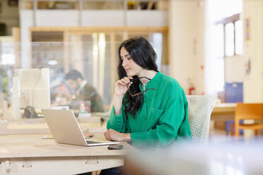 Junge Geschäftsfrau, die auf einen Laptop schaut, während sie am Schreibtisch in einem Coworking-Büro sitzt - EIF00938