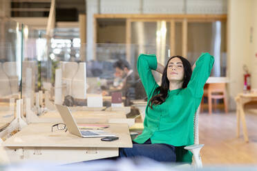 Businesswoman with hands behind head relaxing at desk in coworking office - EIF00940