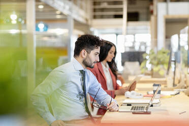 Männlicher Unternehmer, der eine Brille hält, während eine Kollegin in einem Coworking-Büro auf ihren Laptop schaut - EIF00958