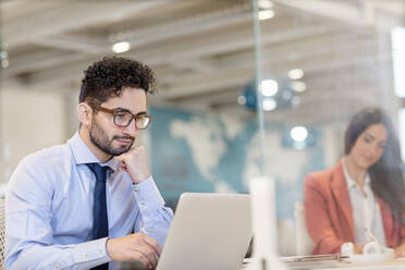 Junger Geschäftsmann mit Brille arbeitet am Laptop mit einer Kollegin im Hintergrund im Büro - EIF00960