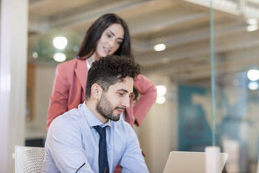 Geschäftsmann, der an einem Laptop arbeitet, während eine Kollegin in einem Coworking-Büro dahinter steht - EIF00969
