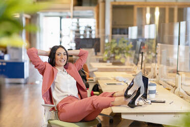 Entspannte weibliche Fachkraft, die lächelnd mit hochgelegten Füßen am Schreibtisch im Büro sitzt - EIF00977