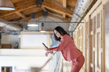 Smiling businesswoman using smart phone while leaning on railing in corridor at office - EIF01001