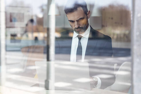 Seriöser Geschäftsmann mit Laptop durch Glasfenster im Büro gesehen - JSRF01507