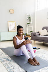 Smiling woman using smart phone on exercise mat at home - GIOF12592