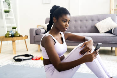 Junge Frau in Sportkleidung benutzt ihr Smartphone, während sie zu Hause sitzt - GIOF12593