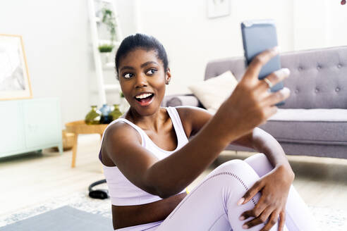 Junge Frau in Sportkleidung, die ein Selfie macht, während sie zu Hause sitzt - GIOF12594