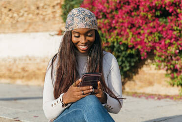 Black female in casual outfit sitting in urban park and chatting on social media via smartphone - ADSF23781