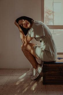 Side view of tender female leaning on hands while sitting with closed eyes on stool in room lit by sunlight - ADSF23830