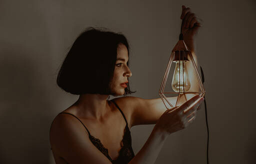Seitenansicht einer zierlichen Frau, die eine beleuchtete Metalllaterne berührt, während sie in einem Zimmer zu Hause sitzt - ADSF23833