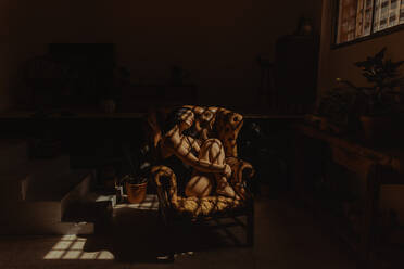 Seitenansicht einer friedlichen Frau im Nachthemd, die in einem von Sonnenlicht beleuchteten Ledersessel sitzt und sich mit geschlossenen Augen entspannt - ADSF23835