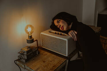 High angle of dreamy female leaning on radio set in retro room and looking at illuminated light bulb - ADSF23841