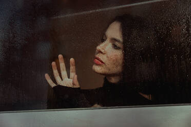 Through glass of thoughtful female standing near window and observing street while looking away - ADSF23847