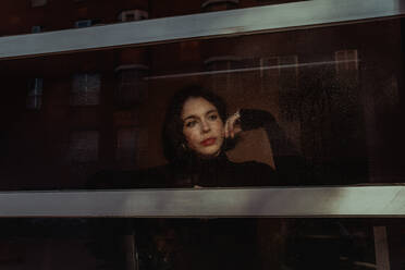 Durch das Glas einer nachdenklichen Frau, die am Fenster steht und die Straße beobachtet, während sie wegschaut - ADSF23848