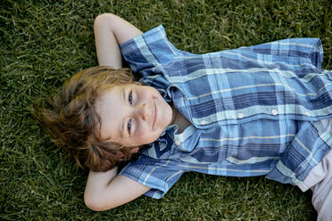 Draufsicht auf ein glückliches kleines Kind im karierten Hemd, das lächelt und in die Kamera schaut, während es mit den Händen hinter dem Kopf auf dem Rasen liegt - ADSF23853