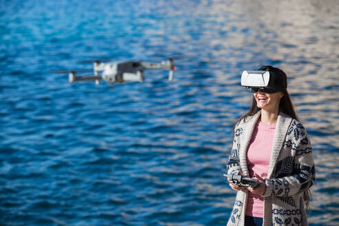 Aufgeregte Frau mit moderner VR-Brille, die eine Drohne mit einer Fernbedienung bedient und virtuelle Realität erlebt, während sie vor einem verschwommenen Meer steht - ADSF23876