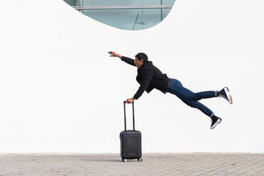 Side view of stylish adult ethnic male traveler with luggage and raised arm flying over walkway while looking forward in town - ADSF23886