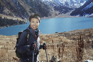 Smiling woman wearing backpack holding hiking poles by lake - AZF00312