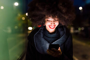 Young woman looking at phone and smiling, illuminated at night - ISF24395