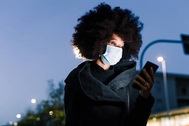 Woman wearing face mask and looking at phone, outdoors at night - ISF24396