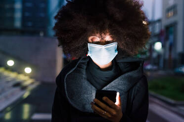 Woman wearing face mask, looking at mobile phone, illuminated at night - ISF24397