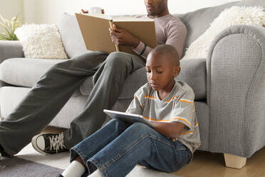 Junge sitzt auf dem Boden und benutzt ein digitales Tablet, Vater liest ein Buch - ISF24544