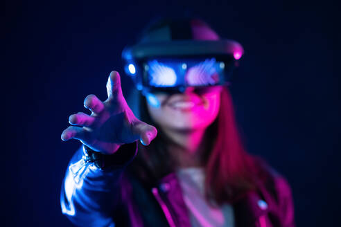 Unrecognizable female with outstretched arm wearing VR headset while exploring virtual reality under blue neon light - ADSF23935