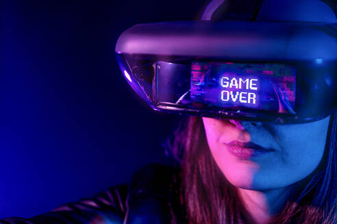 Unrecognizable female wearing modern headset with Game Over inscription while exploring virtual reality in dark room with neon light near wall - ADSF23936