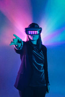Unbekannte Frau mit ausgestrecktem Arm, die ein VR-Headset trägt, während sie die virtuelle Realität unter blauem Neonlicht erkundet - ADSF23943