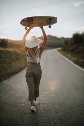 Back view young female in casual wear and summer hat holding cruiser skateboard and looking away while standing on empty asphalt road in rural area at sunset - ADSF23951