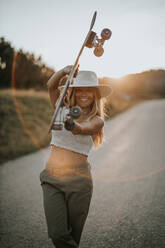 Content young female in casual wear and summer hat holding cruiser skateboard and looking at camera while standing on empty asphalt road in rural area at sunset - ADSF23952
