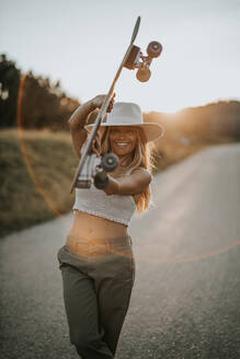 Content young female in casual wear and summer hat holding cruiser skateboard and looking at camera while standing on empty asphalt road in rural area at sunset - ADSF23952