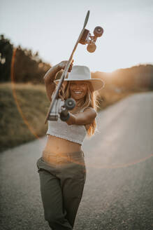 Zufriedene junge Frau in Freizeitkleidung und Sommerhut, die ein Cruiser-Skateboard hält und in die Kamera schaut, während sie auf einer leeren Asphaltstraße in einer ländlichen Gegend bei Sonnenuntergang steht - ADSF23952