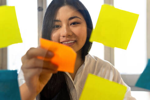 Young glad ethnic female entrepreneur arranging colorful paper stickers on transparent surface in office in daytime - ADSF23954