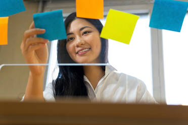Young glad ethnic female entrepreneur arranging colorful paper stickers on transparent surface in office in daytime - ADSF23955