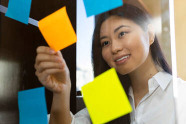 Young glad ethnic female entrepreneur arranging colorful paper stickers on transparent surface in office in daytime - ADSF23956