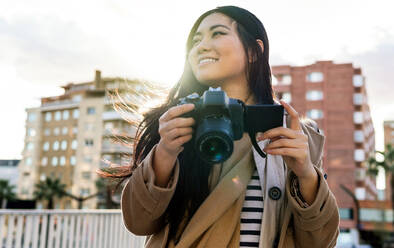 Ethnischer junger glücklicher asiatischer weiblicher Fotograf, der ein Foto mit einer professionellen Fotokamera auf einer städtischen Straße schießt - ADSF23958